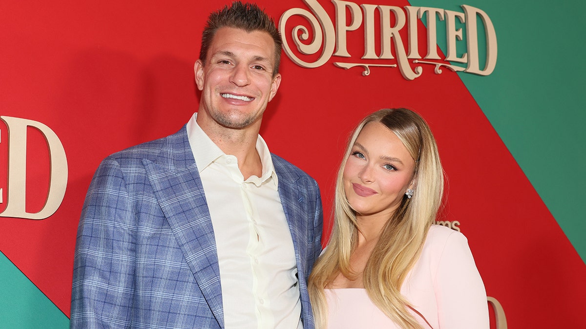 Rob Gronkowski in a blue plaid jacket and white button down smiles next to girlfriend Camille Kostek in a baby pink long-sleeve dress