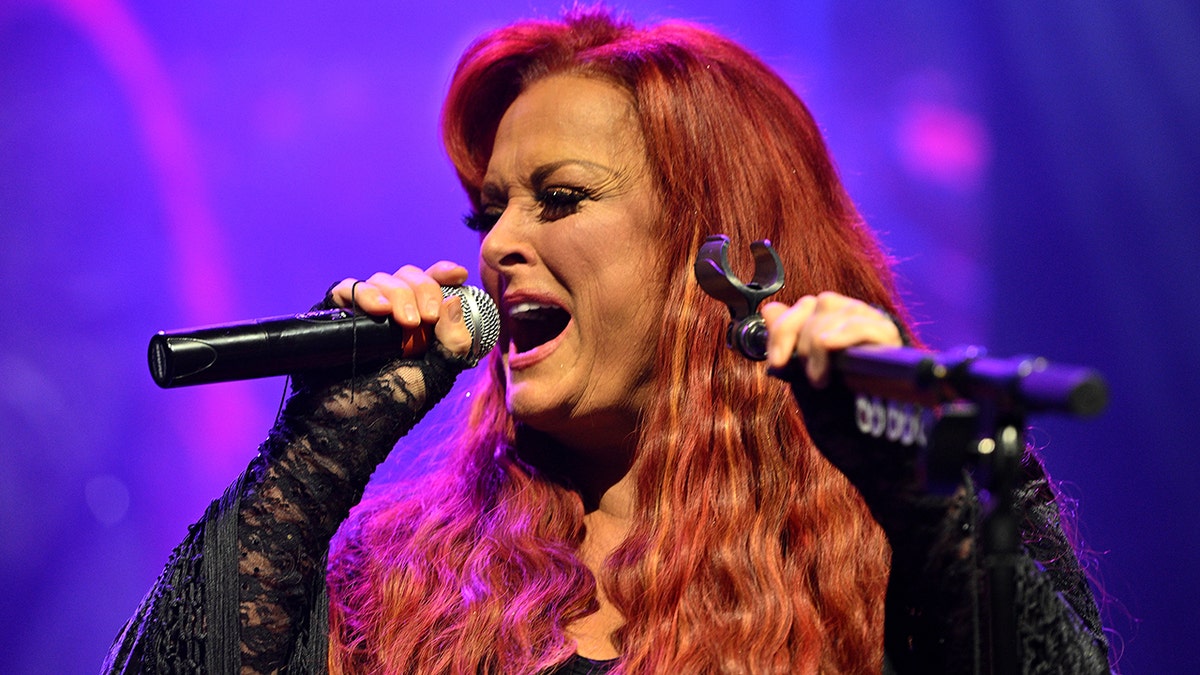 Wynonna Judd with long curly red hair sings into the microphone passionately while on tour in Missouri