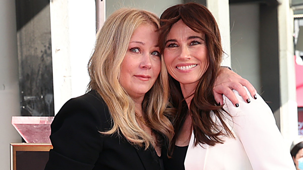 Christina Applegate in black poses on the Hollywood Walk of Fame as she accepts her star and hugs Linda Cardellini, in white smiling