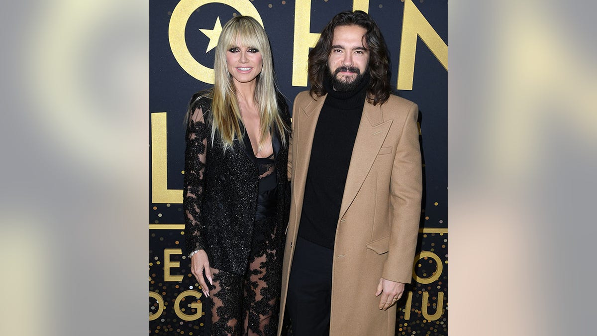 Heidi Klum in a black lace outfit and straightened blonde hair poses on the carpet with husband Tom Kaulitz in a black turtleneck and camel jacket