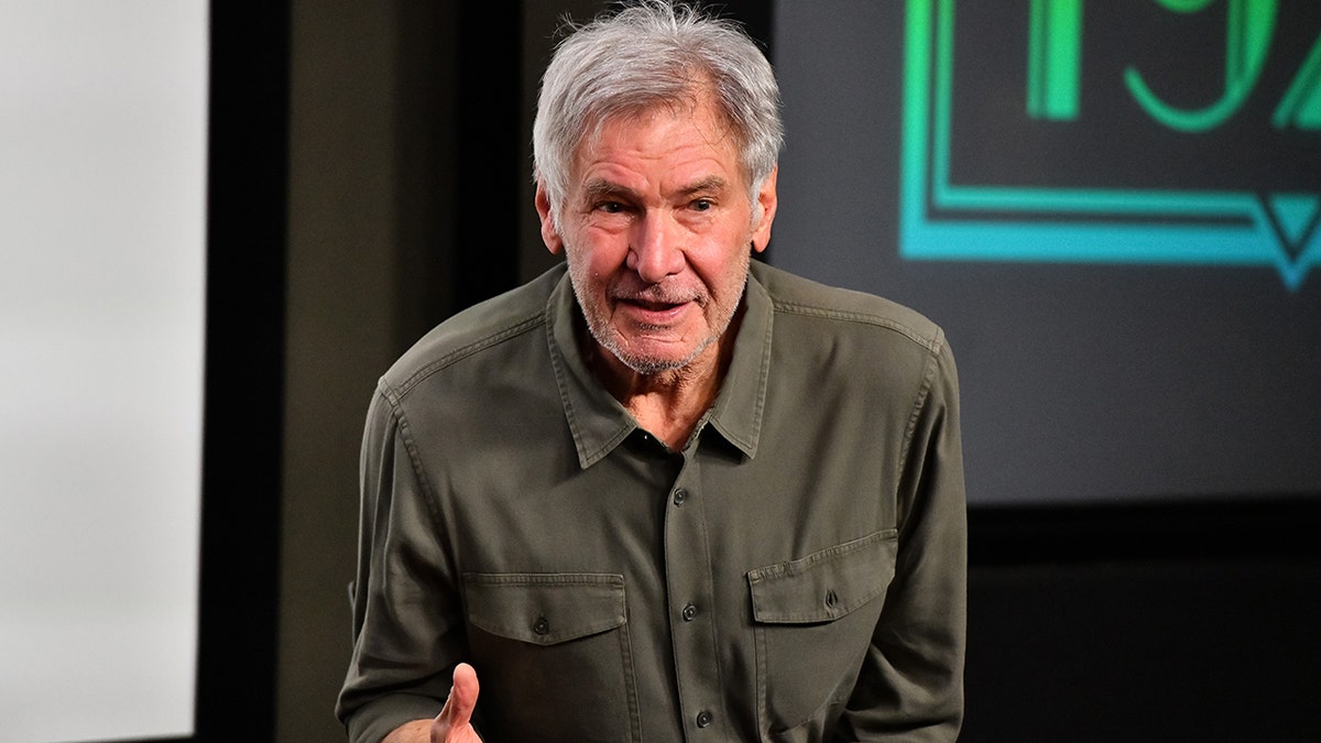 Harrison Ford in an olive green shirt with pockets speaks to a crowd