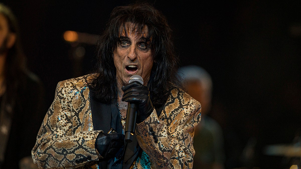 Jeff Beck in a gold and black snakeskin jacket and a thick layer of eyeliner holds up a microphone on stage