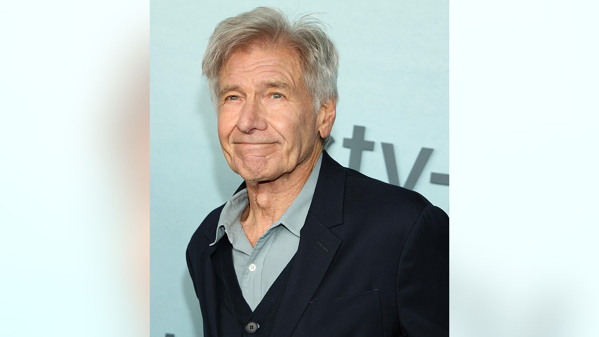 Harrison Ford soft smiles in a light turquoise shirt and black suit jacket on the red carpet