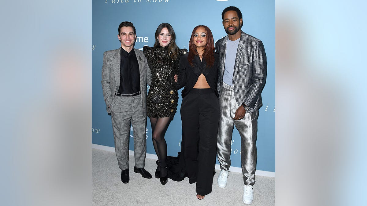 Dave Franco wears a black striped shirt with a checkered black and white suit, next to wife Alison Brie with a sparkly black mini-dress with poofed shoulder pads, next to Kiersey Clemons in a black top that exposes the stomach and black pants next to Jay Ellis in a light blue shirt, metallic pants, and patterned jacket on the red carpet for "Somebody I Used to Know"