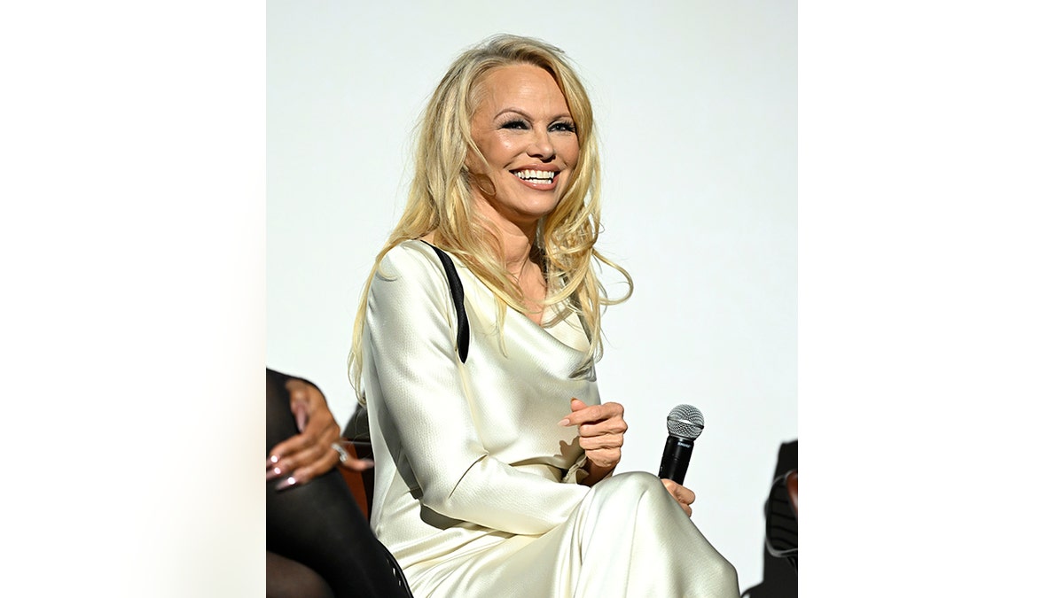 Pamela Anderson smiles on stage in a white silk dress while holding a microphone