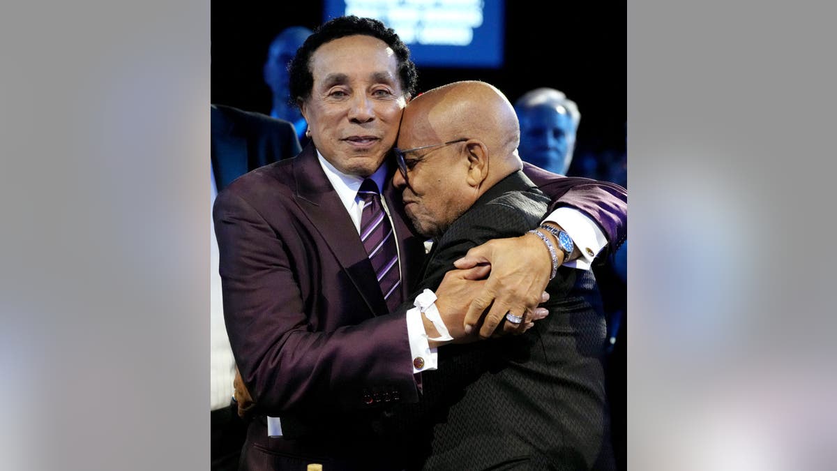 Smokey Robinson in a dark purple suit and purple tie with white stripes is hugged by Berry Gordy in a black suit
