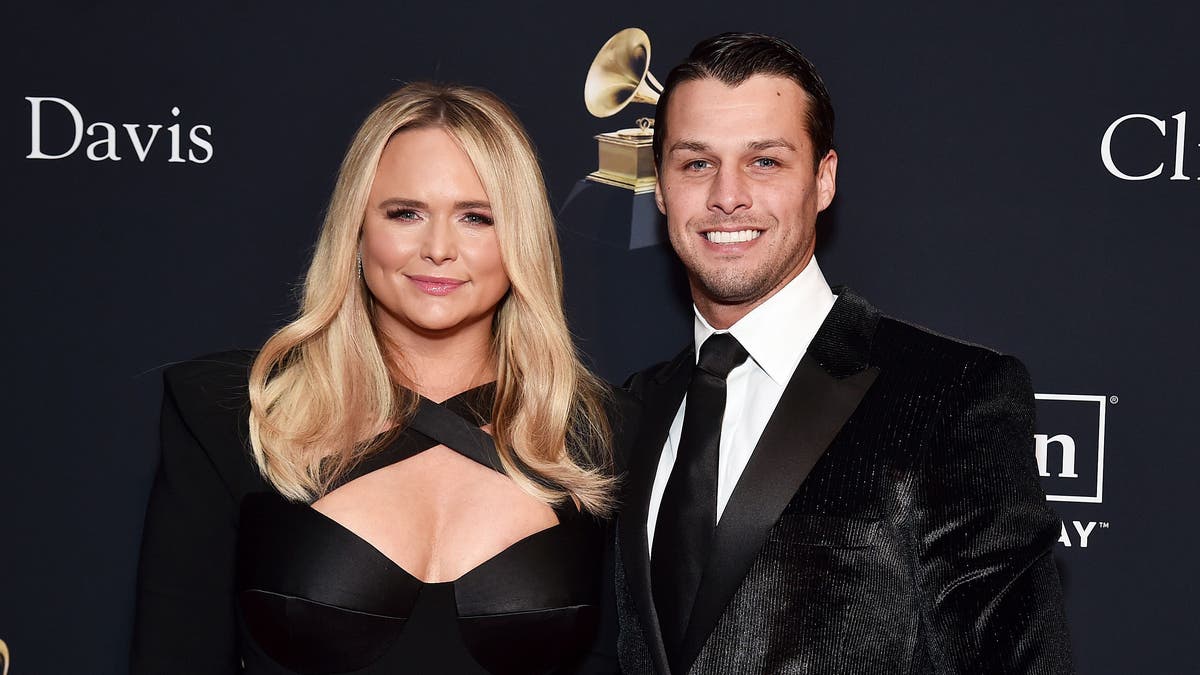 Miranda Lambert soft smiles on the red carpet in a criss-cross cleavage baring black jumpsuit next to her husband Brendan in a shiny black suit