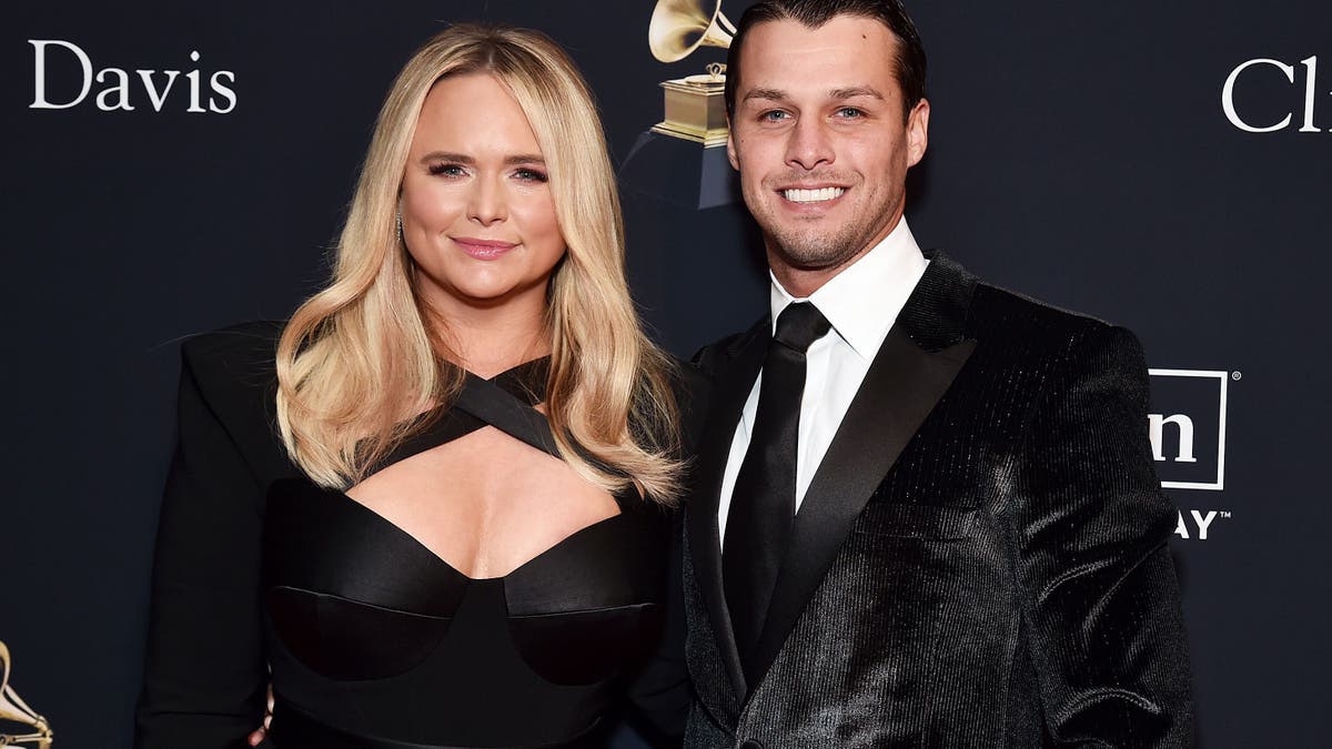 Miranda Lambert soft smiles on the red carpet in a criss-cross cleavage baring black jumpsuit next to her husband Brendan in a shiny black suit