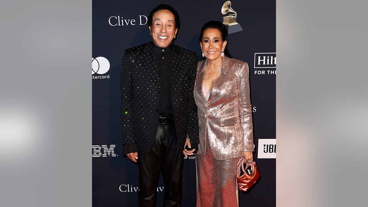 Smokey Robinson in a sparkly black suit jacket and black pants and shirt smiles on the red carpet next to wife Frances Robinson in a rose-gold sparkly dress