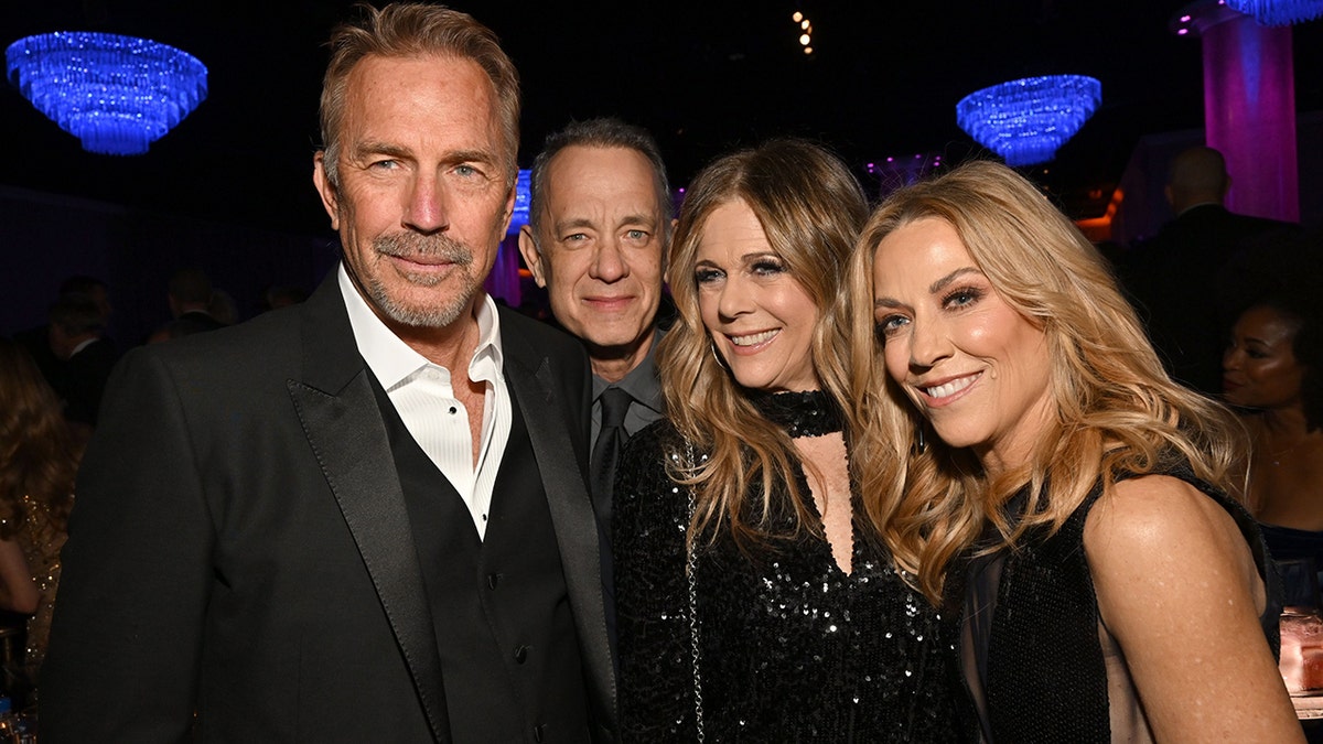 Kevin Costner in a black suit and vest soft smiles for a photo with TomHanks behind his shoulder, Rita Wilson in a sparkly black gown next to him, and Sheryl Crowe in black next to her