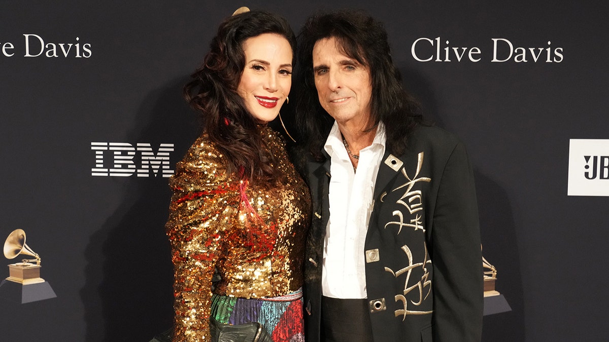 Sheryl Goddard in a sequined gold top with a multi-colored skirt smiles next to husband Alice Cooper wearing a white button down and black jacket with gold 