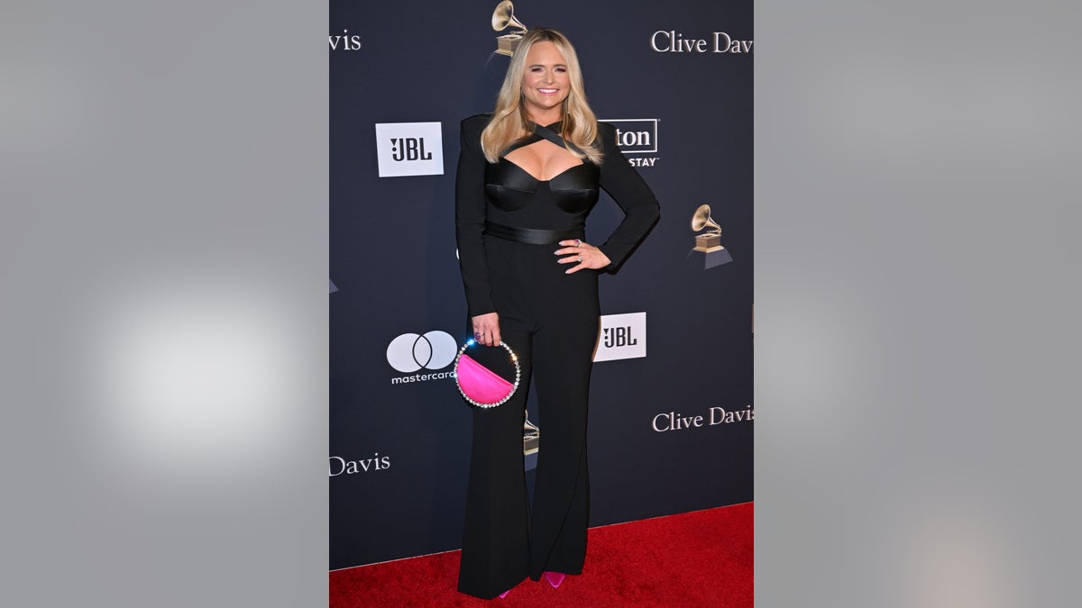 Miranda Lambert in a black criss-cross plunge black jumpsuit poses on the red carpet with a hot-pink bag with sparkles on the handle and around the outline of the bag