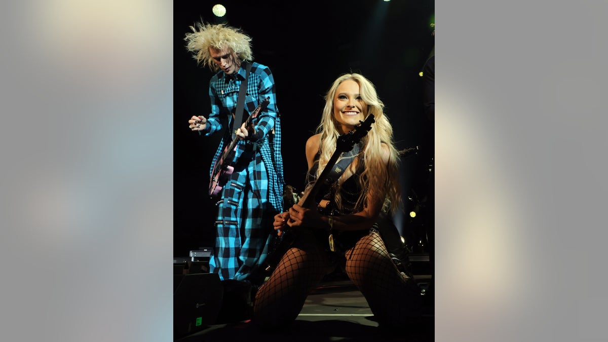 Machine Gun Kelly plays the guitar while on stage in a turquoise and black checkered suit and Sophie Lloyd is on her knees in a black outfit and black fishnet stockings while playing the guitar