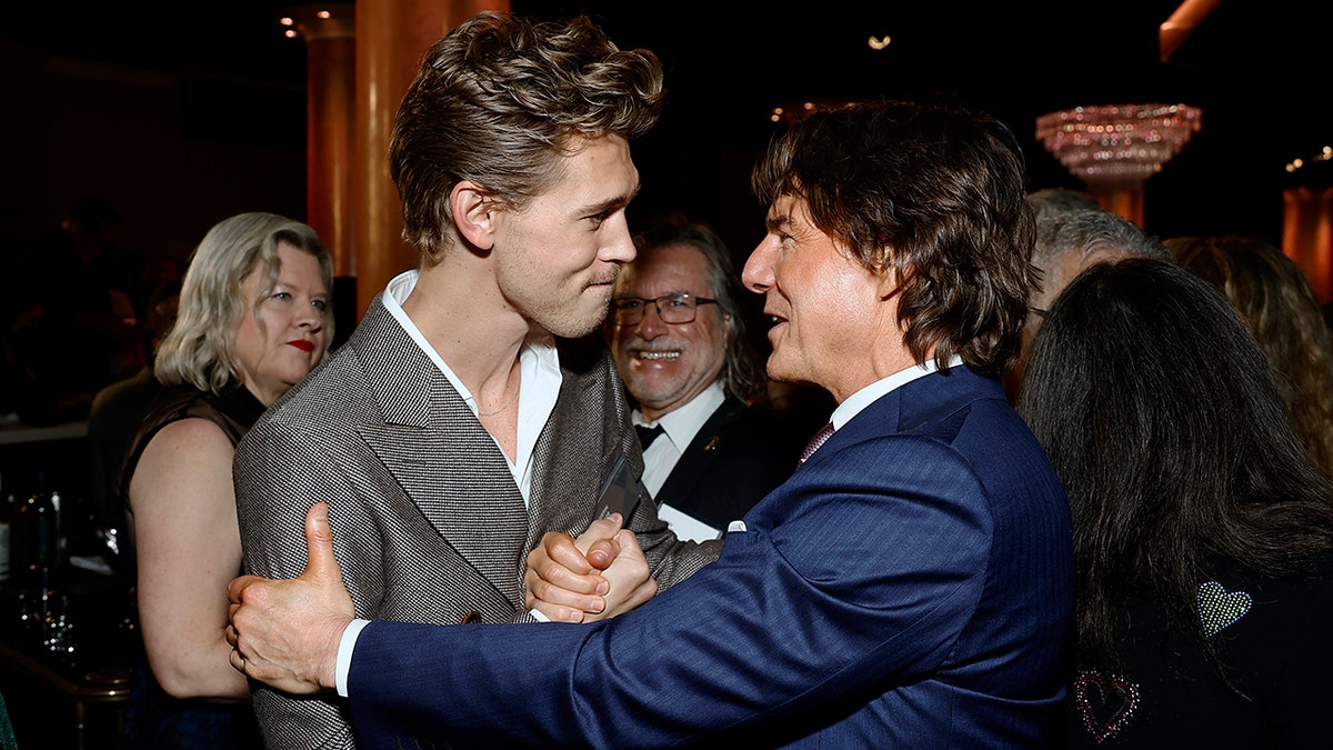 Austin Butler in a grey jacket holds hands with Tom Cruise in a blue jacket who has his arm on Butler's forearm