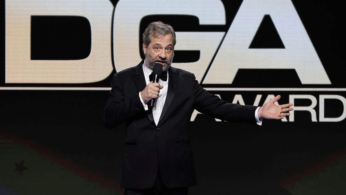 Judd Apatow in a black tuxedo stands on the stage holding a microphone with one arm extended at the DGA Awards