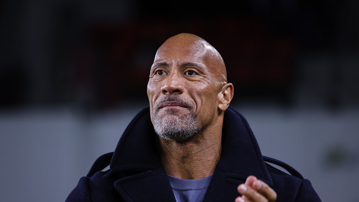 Dwayne Johnson with some grey facial hair claps his hands while watching an XFL game wearing a black pea-coat and light blue shirt