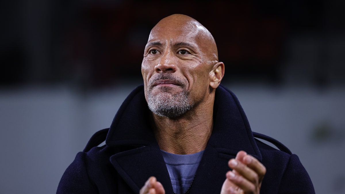 Dwayne Johnson with some grey facial hair claps his hands while watching an XFL game wearing a black pea-coat and light blue shirt