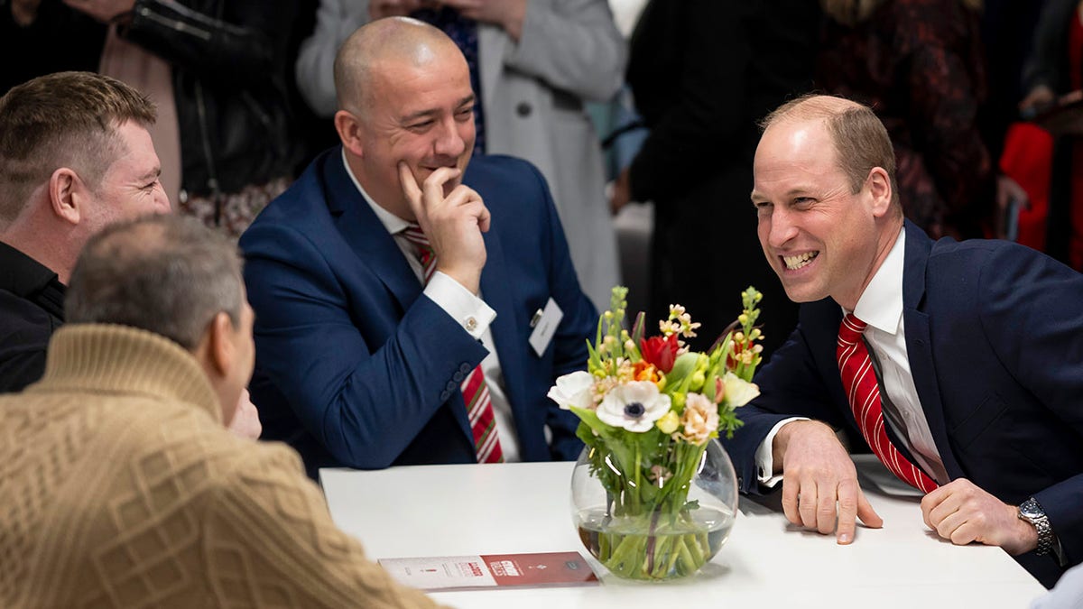 Prince William laughs at a table with a flower bouquet as he enjoys a conversation with injured rugby players in Cardiff