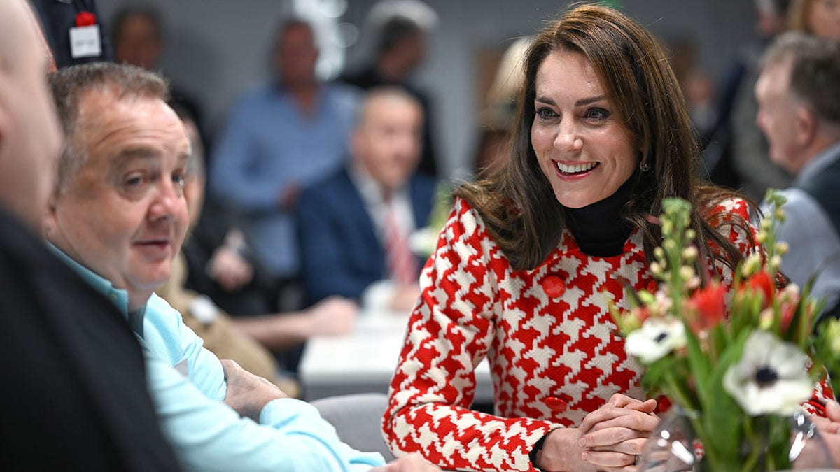 Kate Middleton in a red and white patterned dress sits at a table and beams as she speaks with injured rugby players