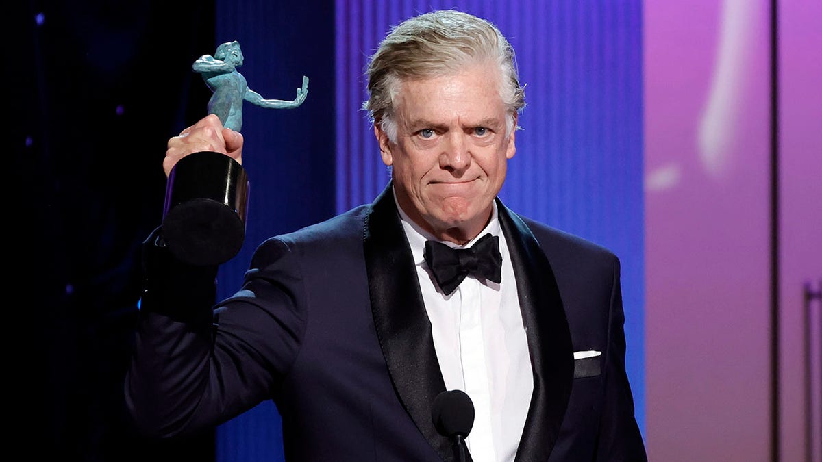 Christopher McDonald in a navy tuxedo with black trim holds up the blue SAG Award given to Jean Smart while accepting it on her behalf
