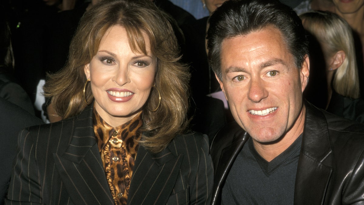 Raquel Welch smiling in a dark coat and an animal printed blouse next to her husband wearing all black.