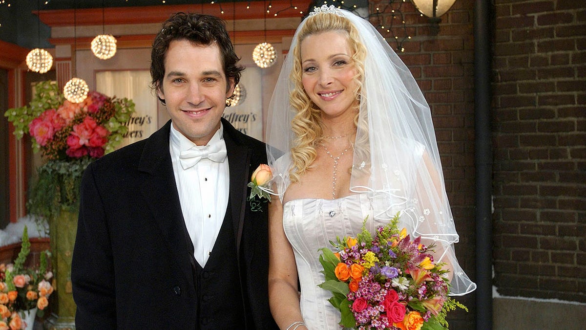 Paul Rudd wears a black tuxedo and white bowtie as Mike Hannigan in "Friends" marrying Lisa Kudrow's character of Phoebe Buffay in a wedding dress and holding a colorful flower arrangement