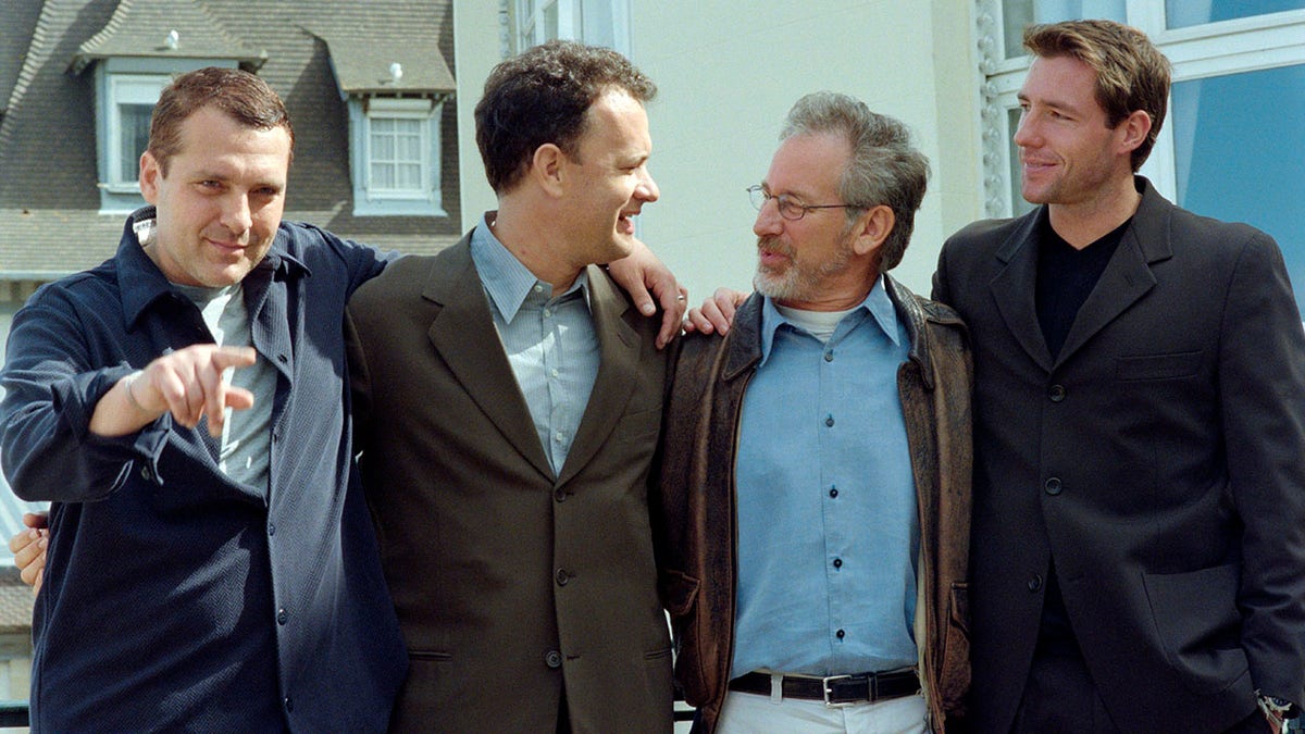 Tom Sizemore points at the camera while posing for a picture with Tom Hanks in a brown jacket who is looking at Steven Spielberg in a bright blue shirt and leather jacket next to actor Edward Burns in a dark suit with buttons in 1998