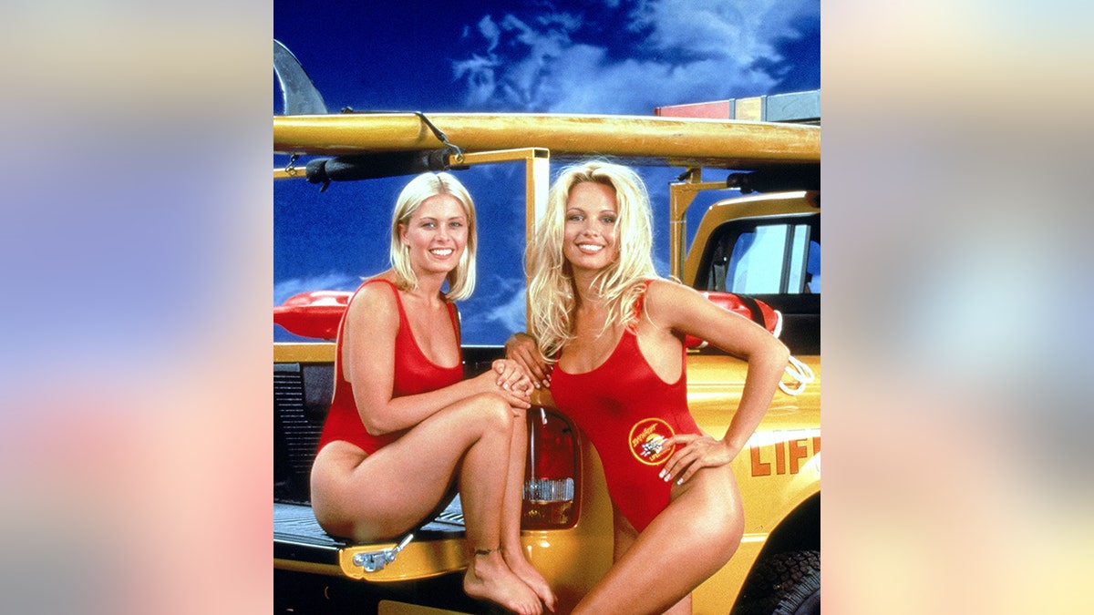 Pamela Anderson and Nicole Eggert pose for a photo in front of a yellow lifeguard truck wearing their red bathing suits from "Baywatch"