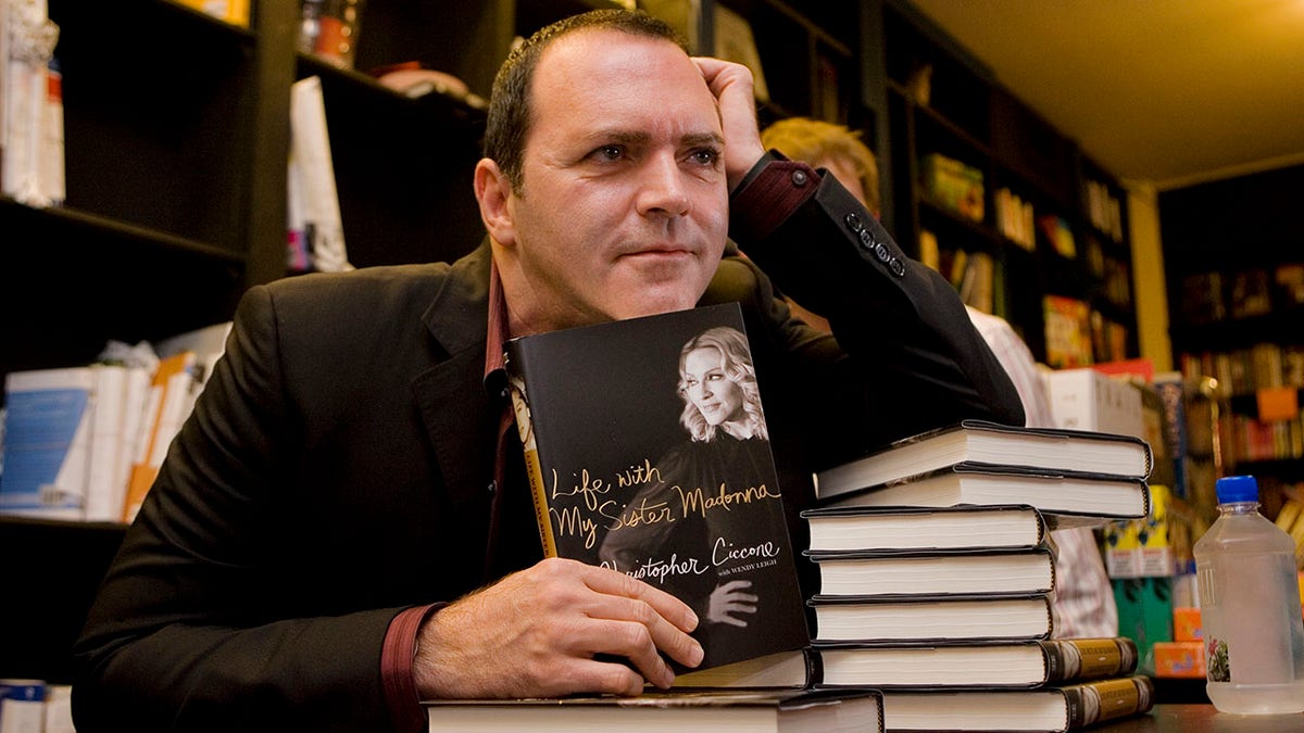 Madonna's brother Anthony Ciccone holds up his book with one hand and puts his other hand to his head, in a black suit