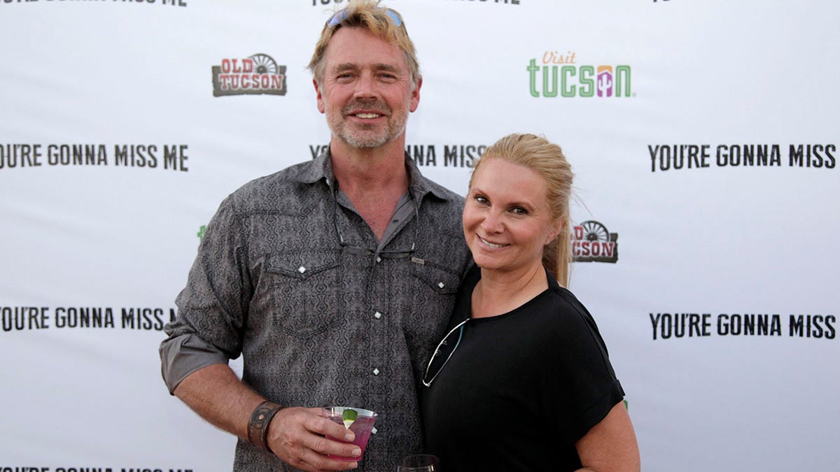 John Schneider in a grey printed button down holds a drink and smiles with his wife Alicia Allain in a black top on the red carpet