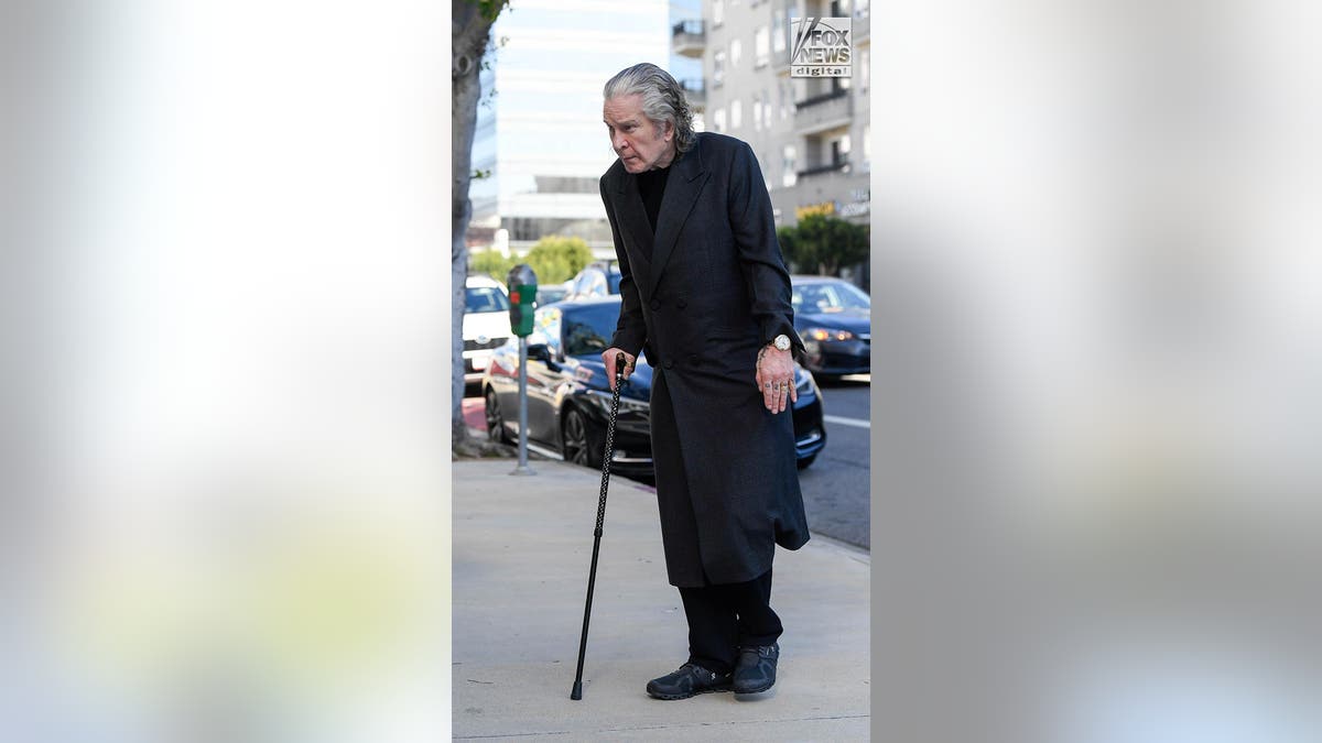 Ozzy Osbourne wearing a black coat over dark clothes walks with a cane