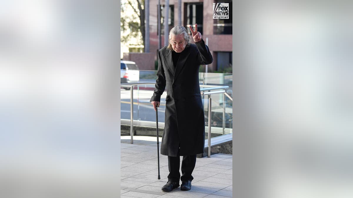Ozzy Osbourne wearing a black coat over dark clothes walks with a cane as he gives the two-finger peace salute