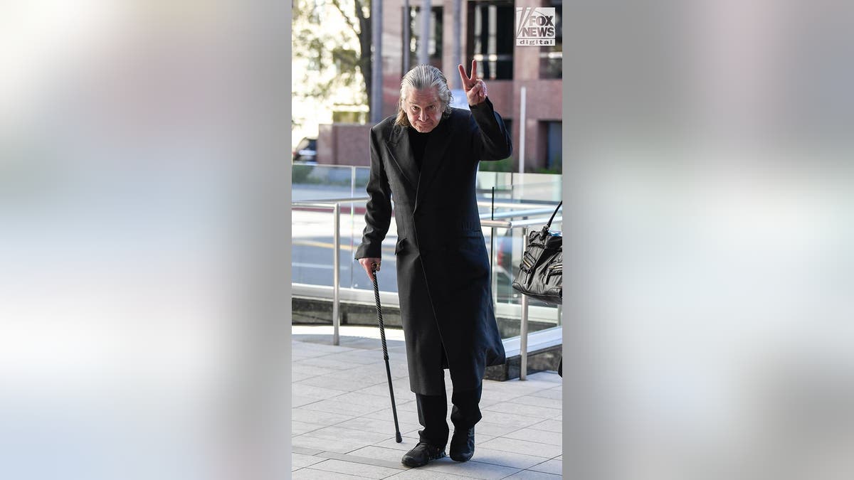 Ozzy Osbourne wearing a black coat over dark clothes walks with a cane as he gives the two-finger peace salute