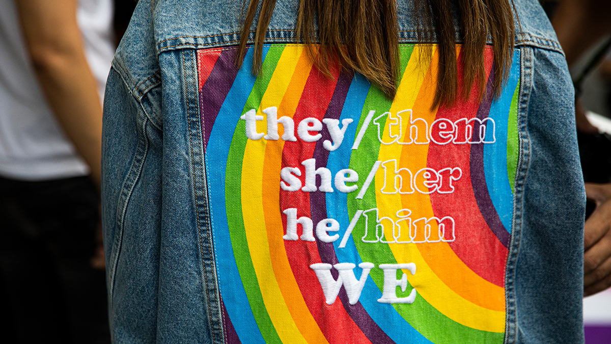 pronouns embroidered on top of rainbow design on back of jean jacket