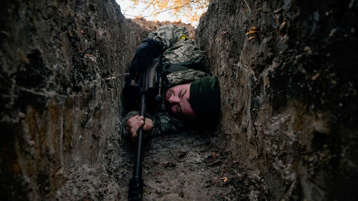 A man wearing military gear hides in a trench.