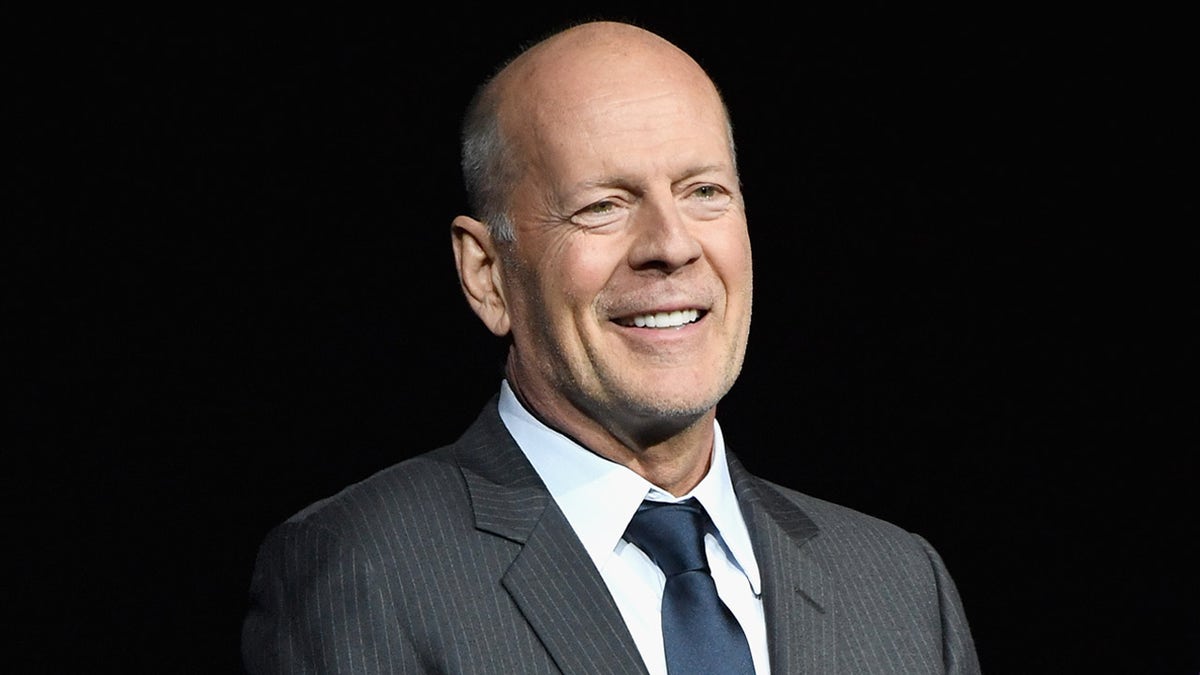 Bruce Willis smiles on stage in a grey suit and navy tie
