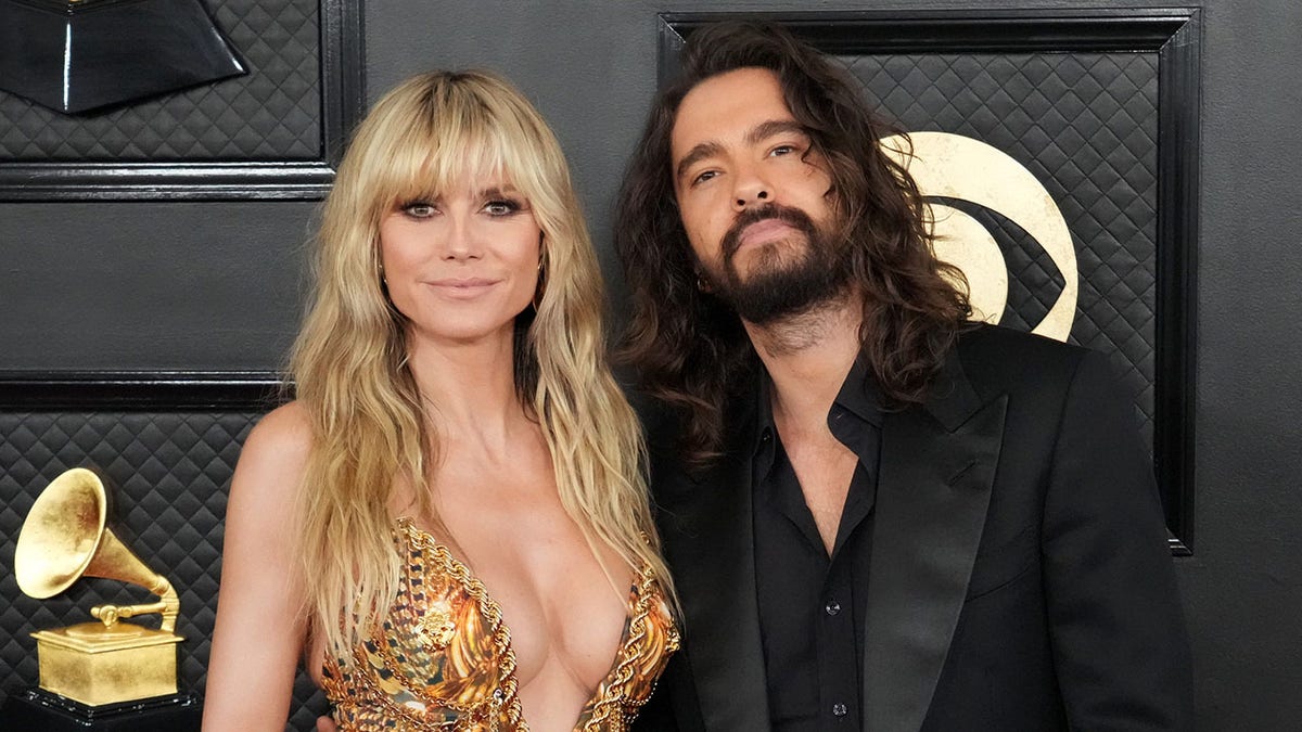 Heidi Klum in a gold low plunging sequin dress poses with husband Tom Kaulitz in a black suit and shirt