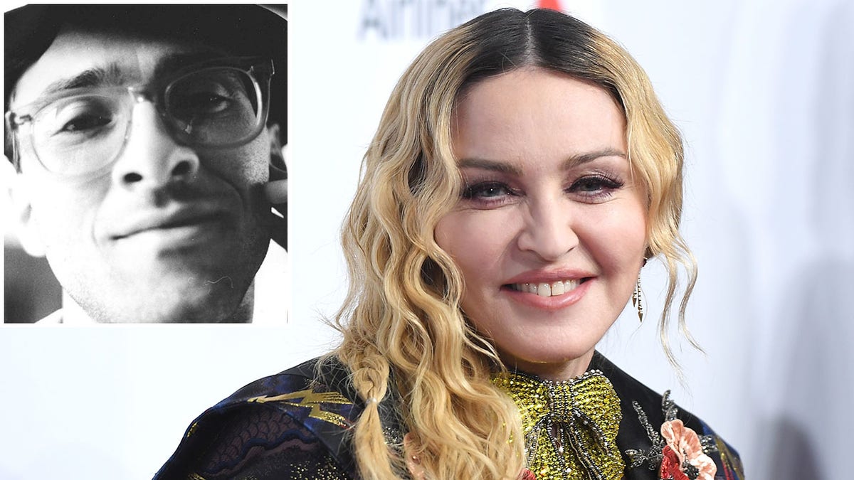 Madonna with curly blonde hair smiles on the red carpet in a multi-colored dress with jewels inset a black-and-white photo of her brother Anthony Ciccone with circular glasses