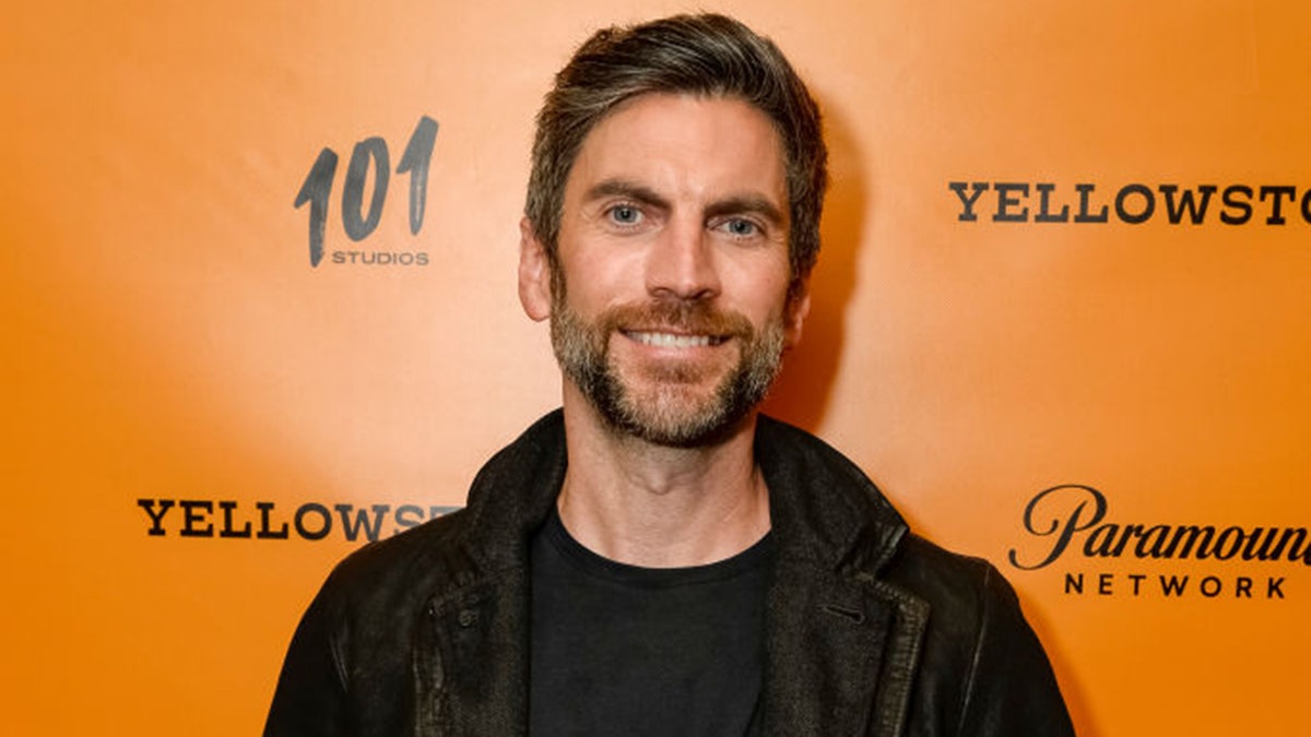 Wes Bentley in black smiles in front of an orange backdrop for a "Yellowstone" premiere