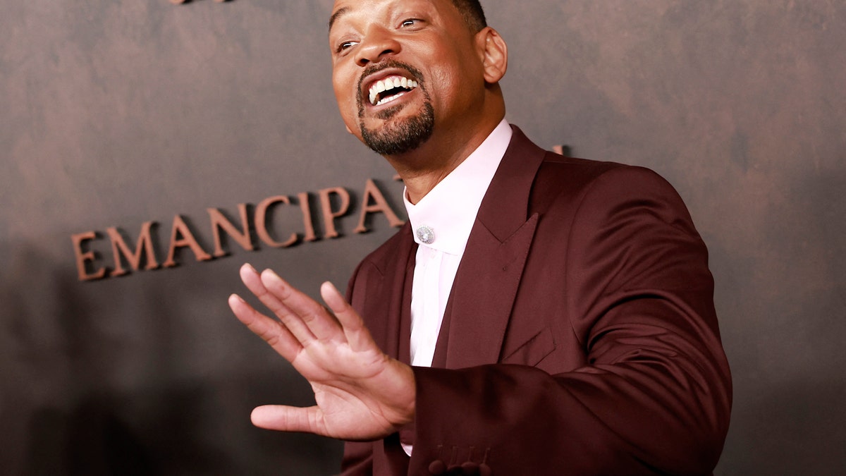 Will Smith smiles and holds up his hand at an event