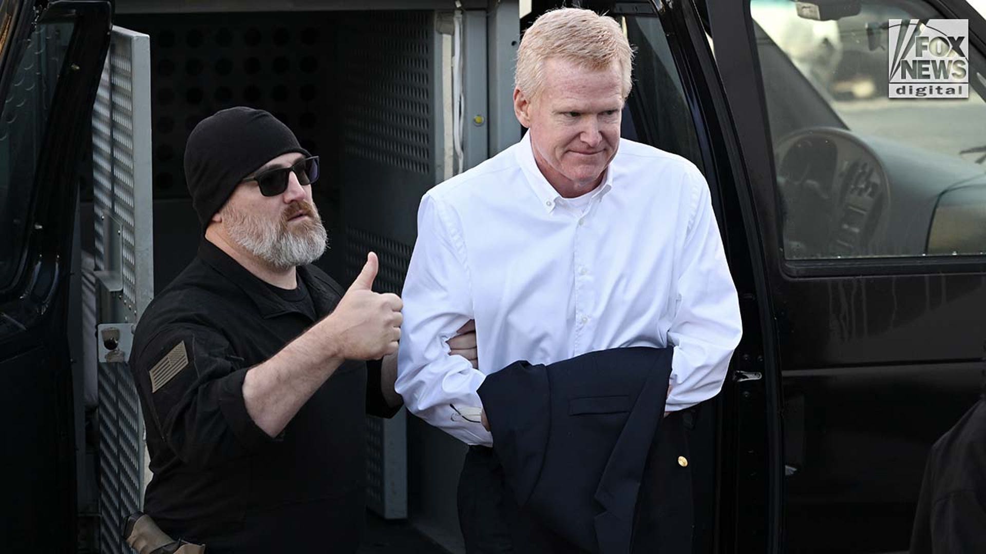 A man in a white button-down exits a police vehicle surrounded by officers.