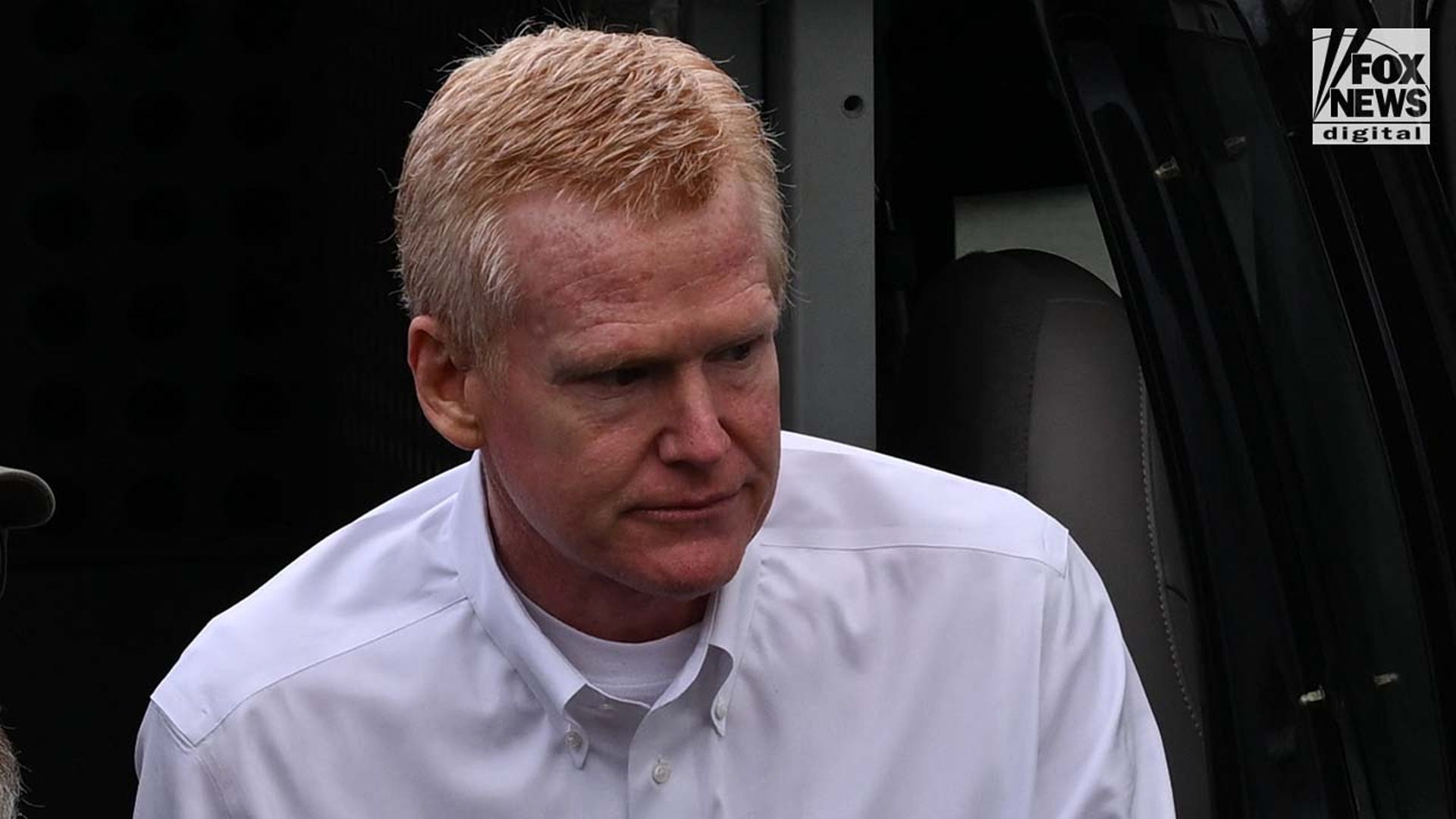 A man wearing a white, dress shirt is escorted out of a police vehicle by armed officers.