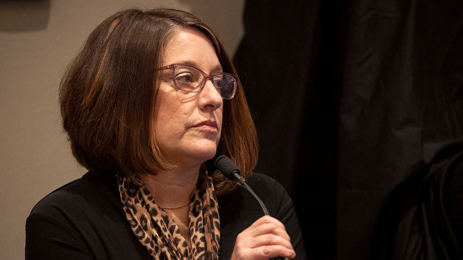 A woman speaks into a microphone while on the witness stand inside a courtroom.