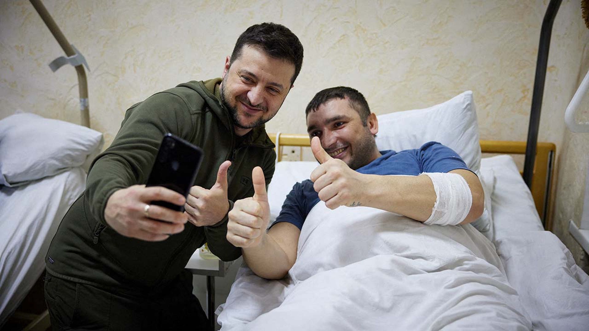 A man in a hospital bed smiles and gives a thumbs up with another man.