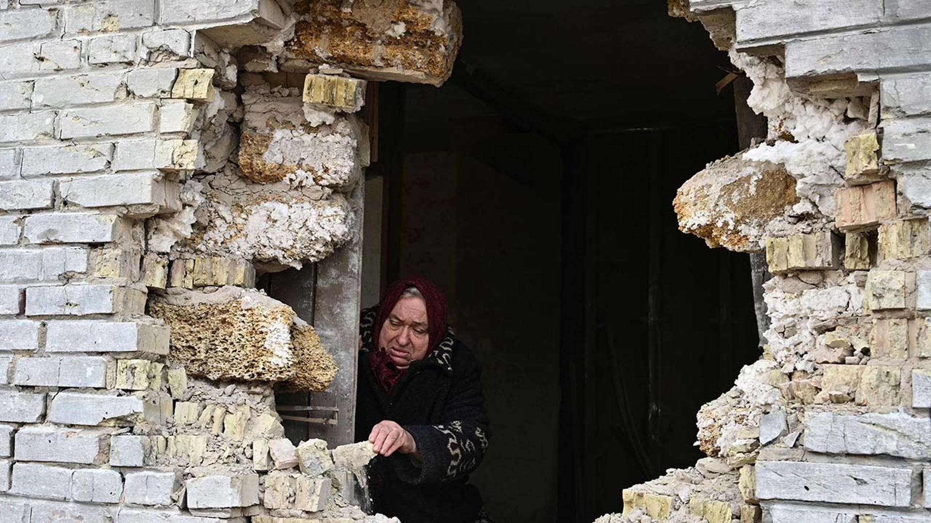 A woman peers out of a large hole in the side of her house.