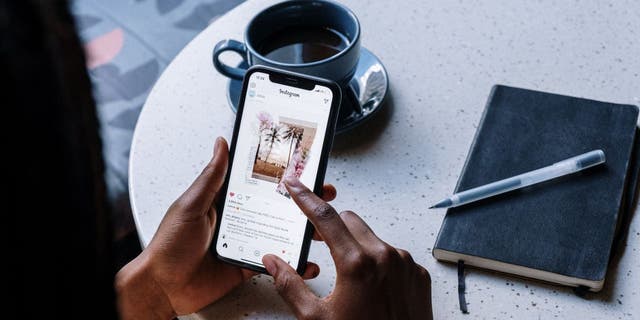 A person views an Instagram post on their phone as they sit at a table with a journal and cup of coffee. 