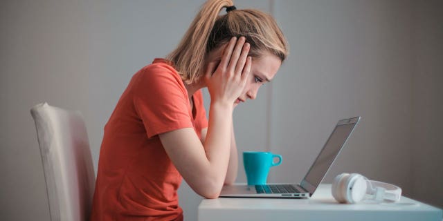 Woman in an orange shirt sitting at the table, staring at her laptop.