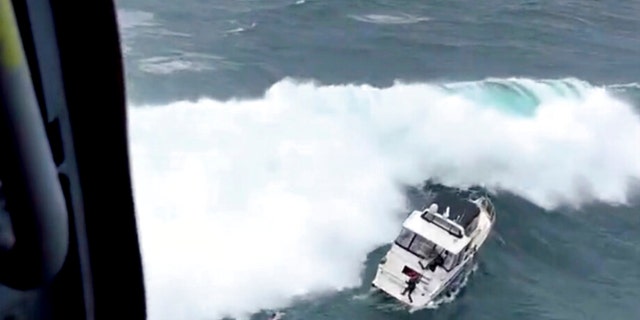 In this photo provided by the U.S. Coast Guard Pacific Northwest, a Coast Guard rescue swimmer reaches a boat right before a giant wave rolled the craft at the mouth of the Columbia River in Oregon on Friday, Feb. 3, 2023. The newly minted Coast Guard rescue swimmer saved the life of a man who was piloting the yacht. 