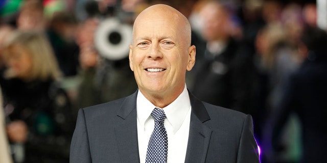 Bruce Willis smiles on the red carpet in a black suit, white button down and blue patterned tie