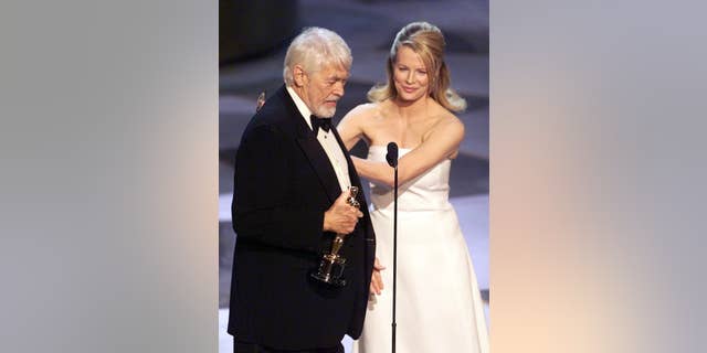 James Coburn holds up his Oscar during the 71st Academy Awards. Presenting the Oscar is Kim Basinger.